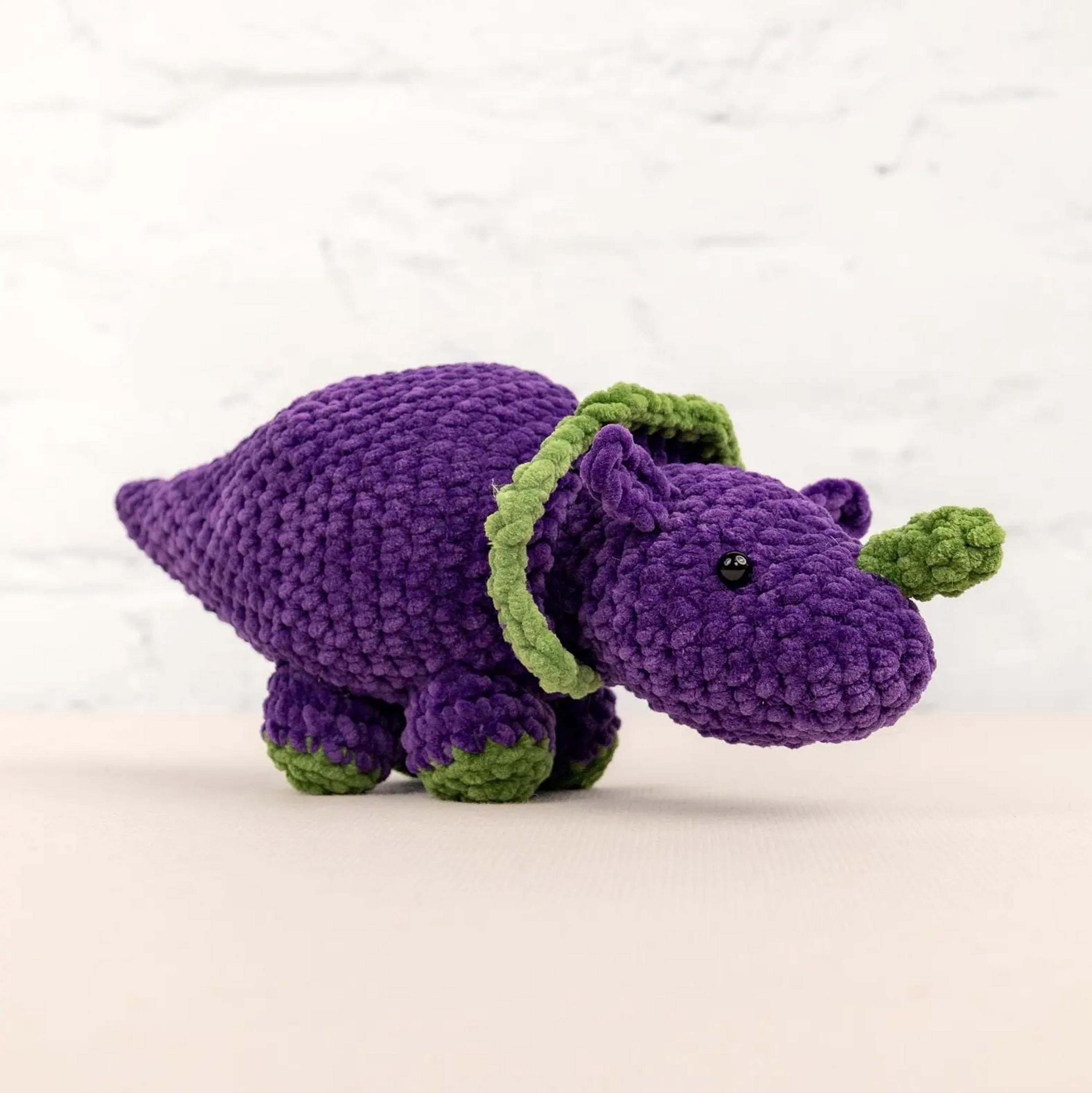 A purple crocheted toy dinosaur with green accents is sitting on a white surface against a white background.